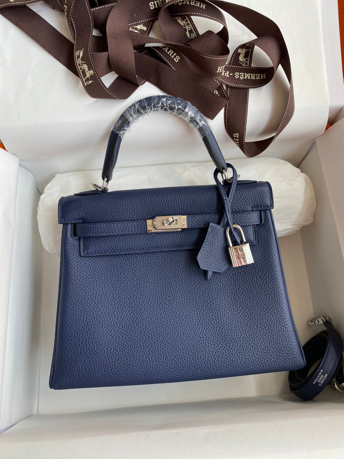 Blue handbag with silver hardware on a white cushion, surrounded by Hermes packaging.