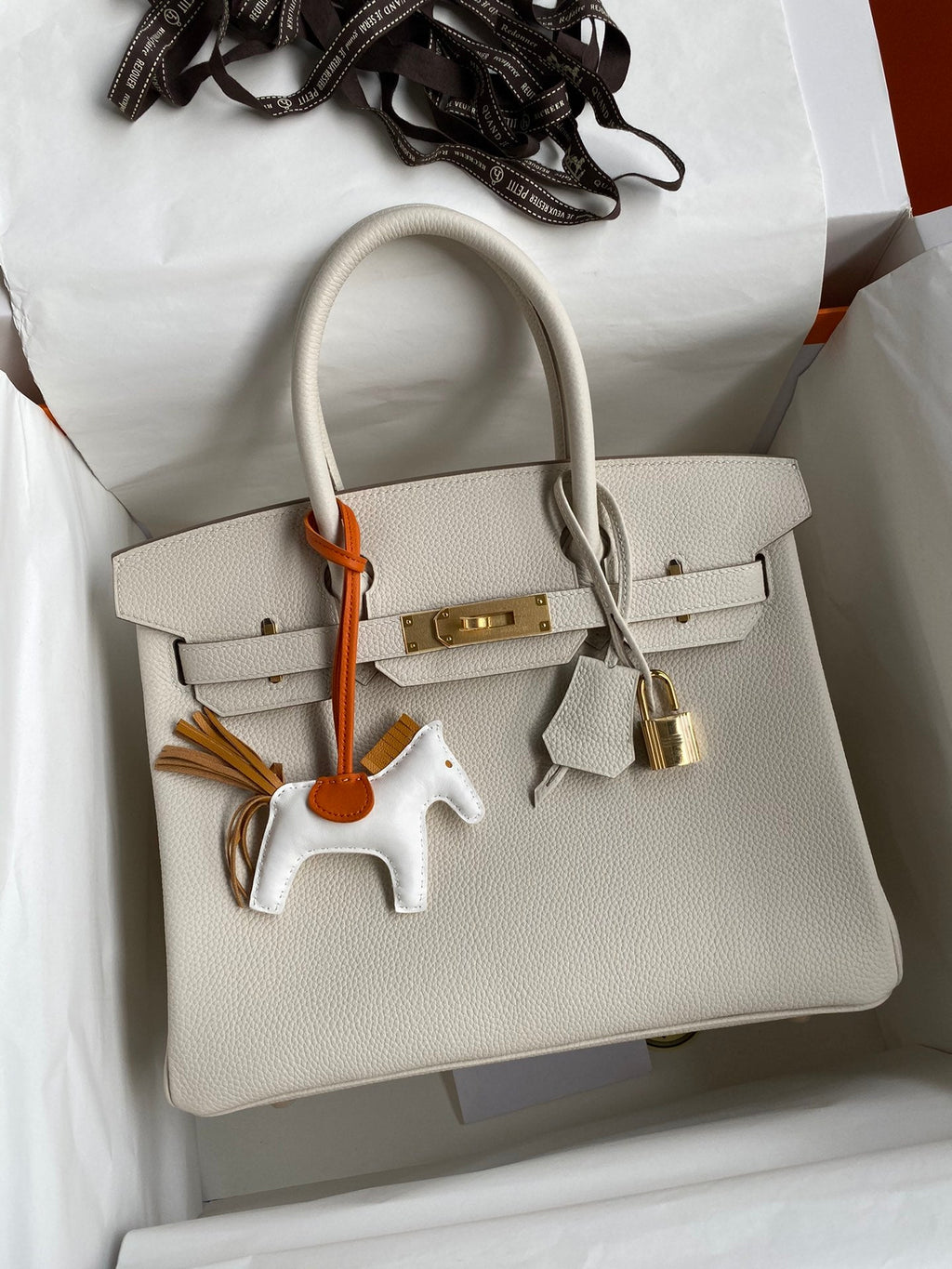 Beige handbag with gold hardware and a horse keychain on a white surface