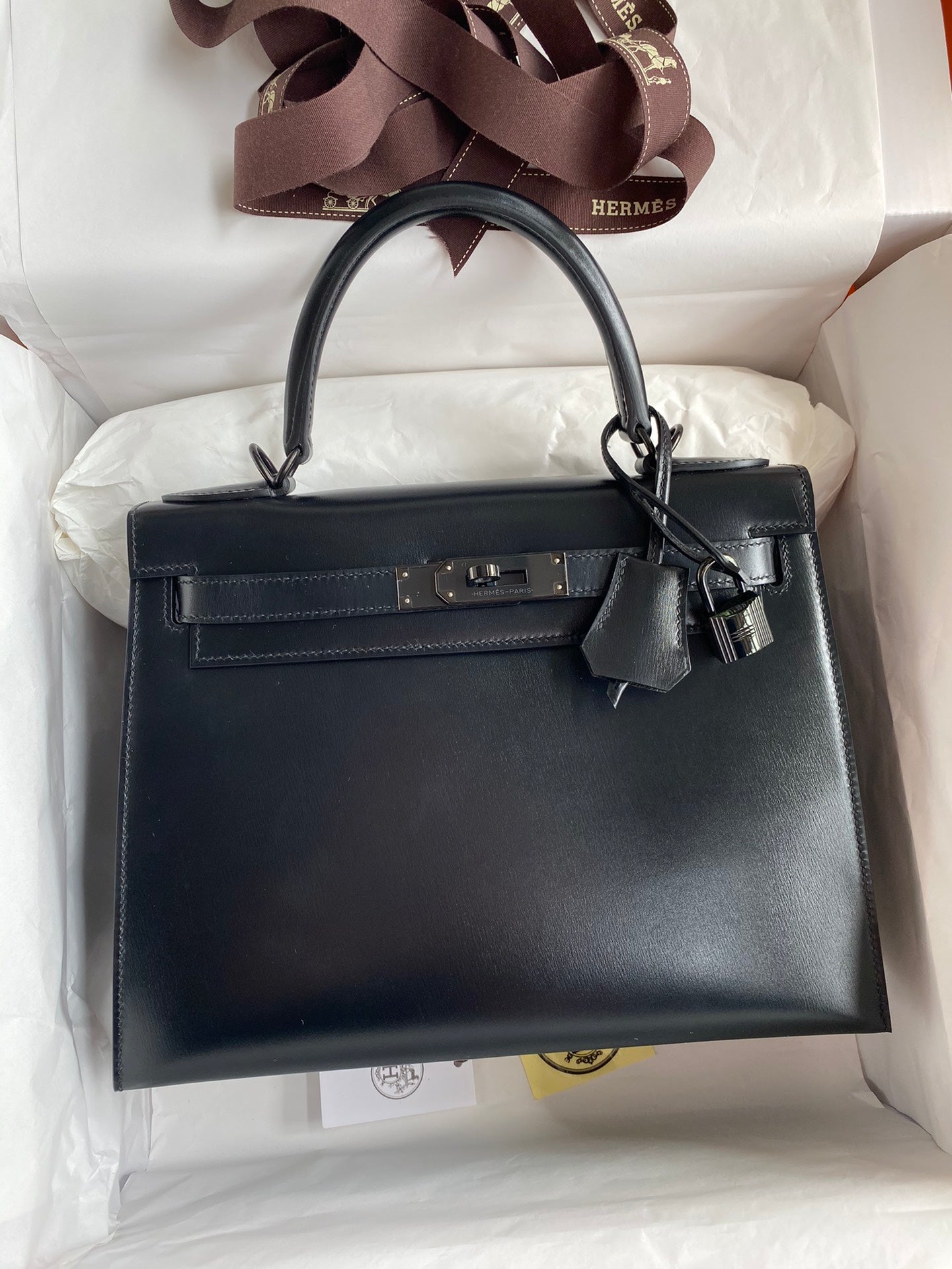 Black handbag with gold hardware in a white box, featuring Hermès branding.