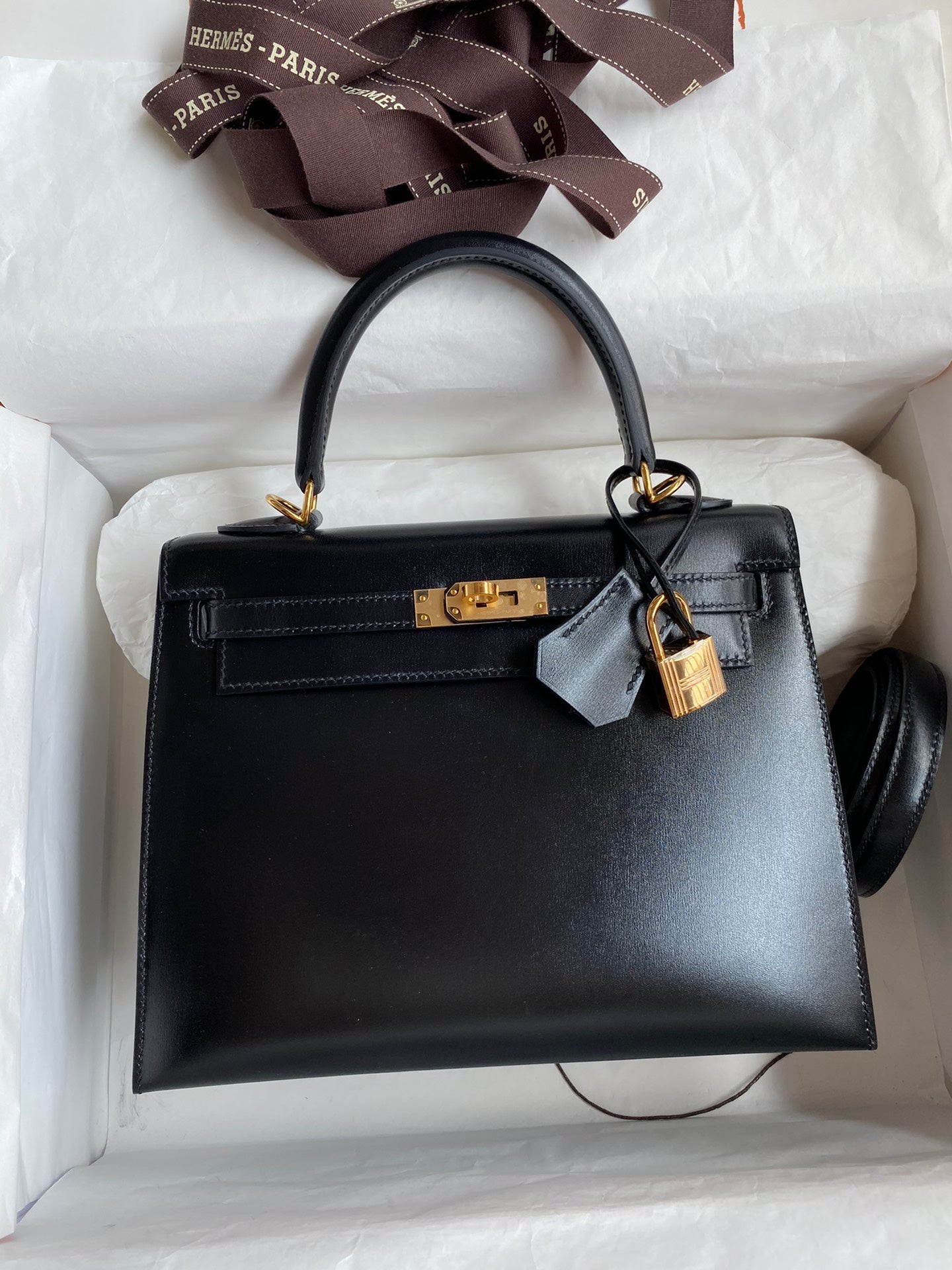 Black handbag with gold hardware and a lock, placed in an open white box.
